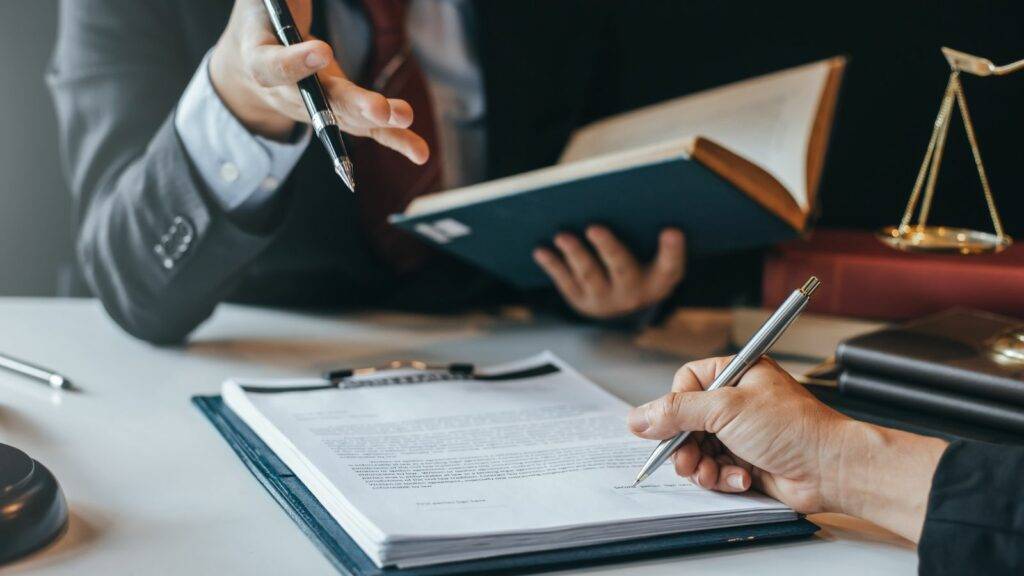 Business people signing legal documents during commercial dispute resolution, with gavel and scales of justice on table.